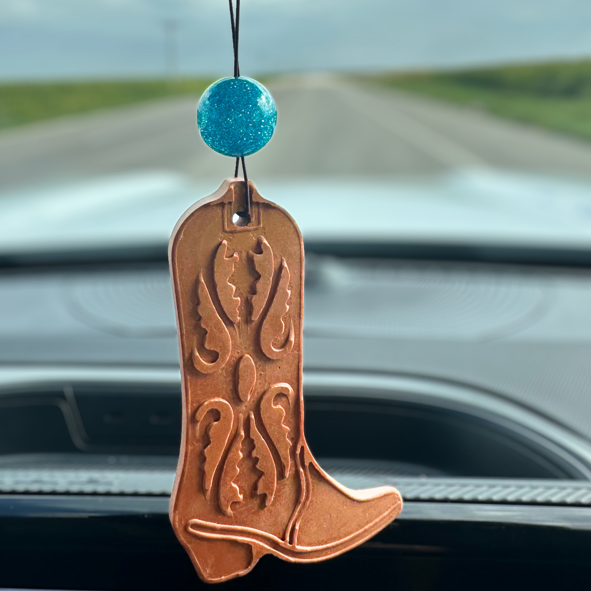 Copper colored cowboy boot-shaped stone car air freshener with a sparkling turquois bead hanging from rearview mirror.