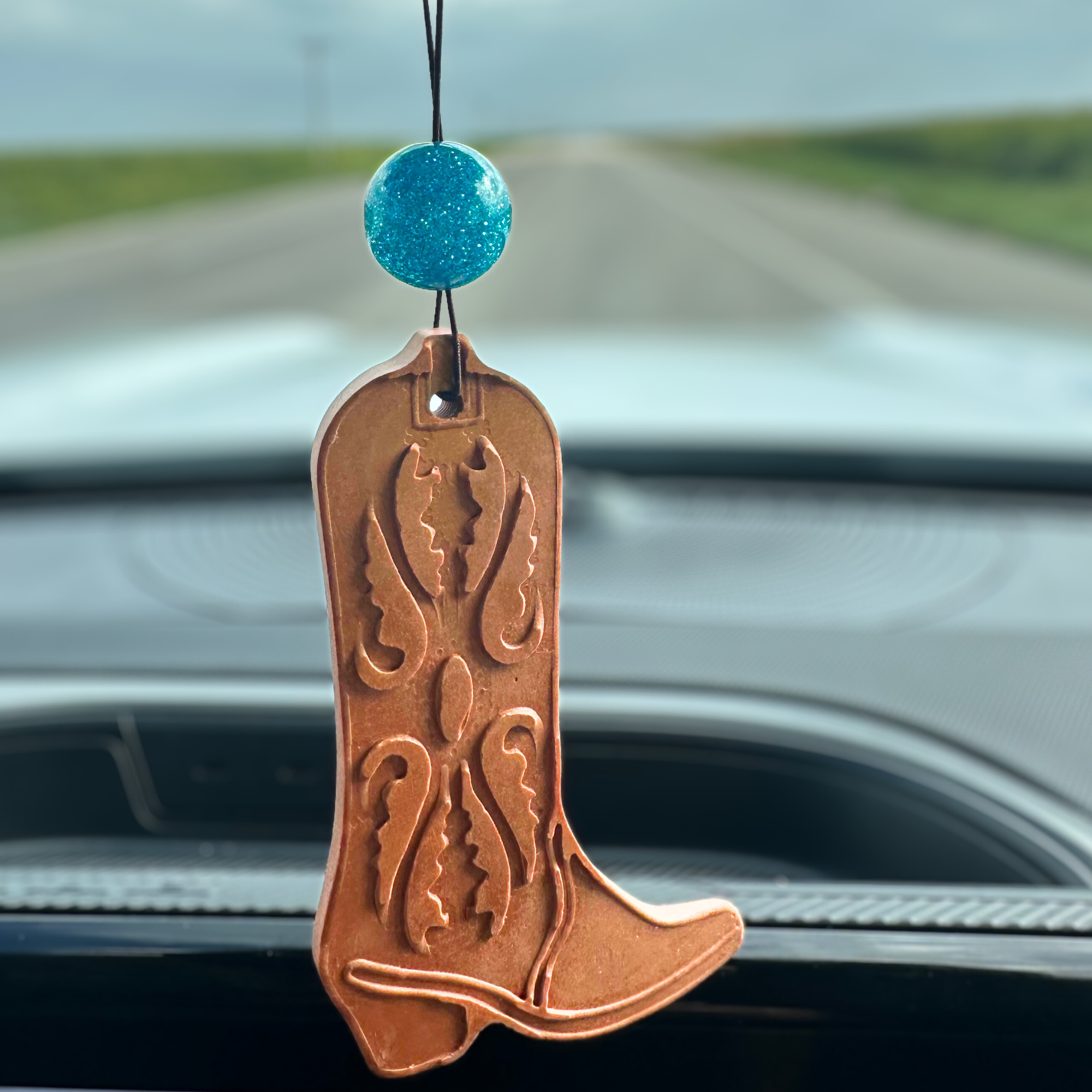 Copper colored cowboy boot-shaped stone car air freshener with a sparkling turquois bead hanging from rearview mirror.