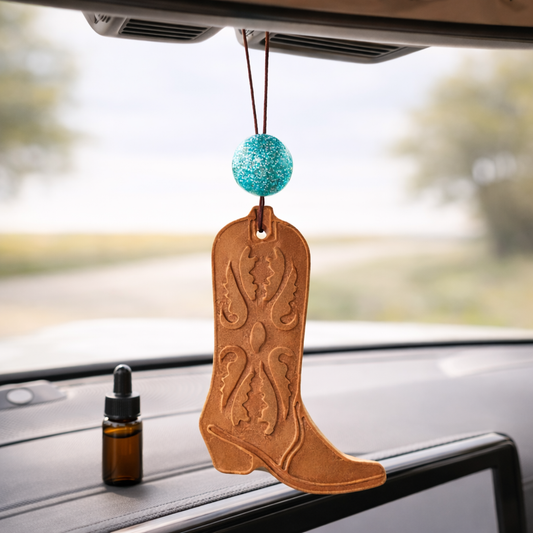 Cowboy boot-shaped car air freshener hanging from a car's rearview mirror with a dropper bottle on a car dashboard.