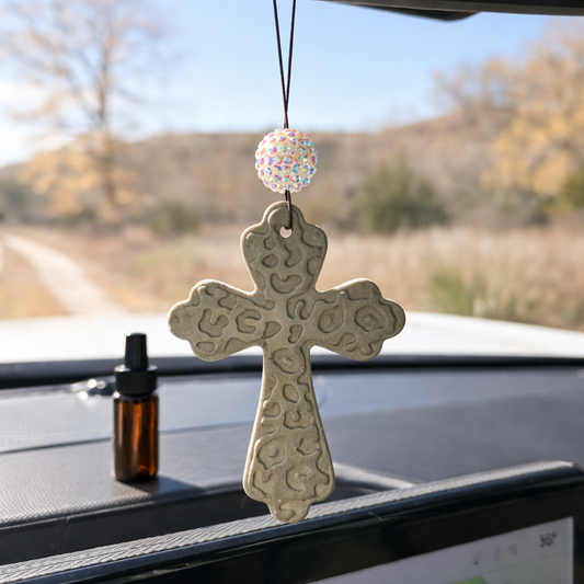 Decorative cross hanging from a car's rearview mirror with a blurred outdoor background.
